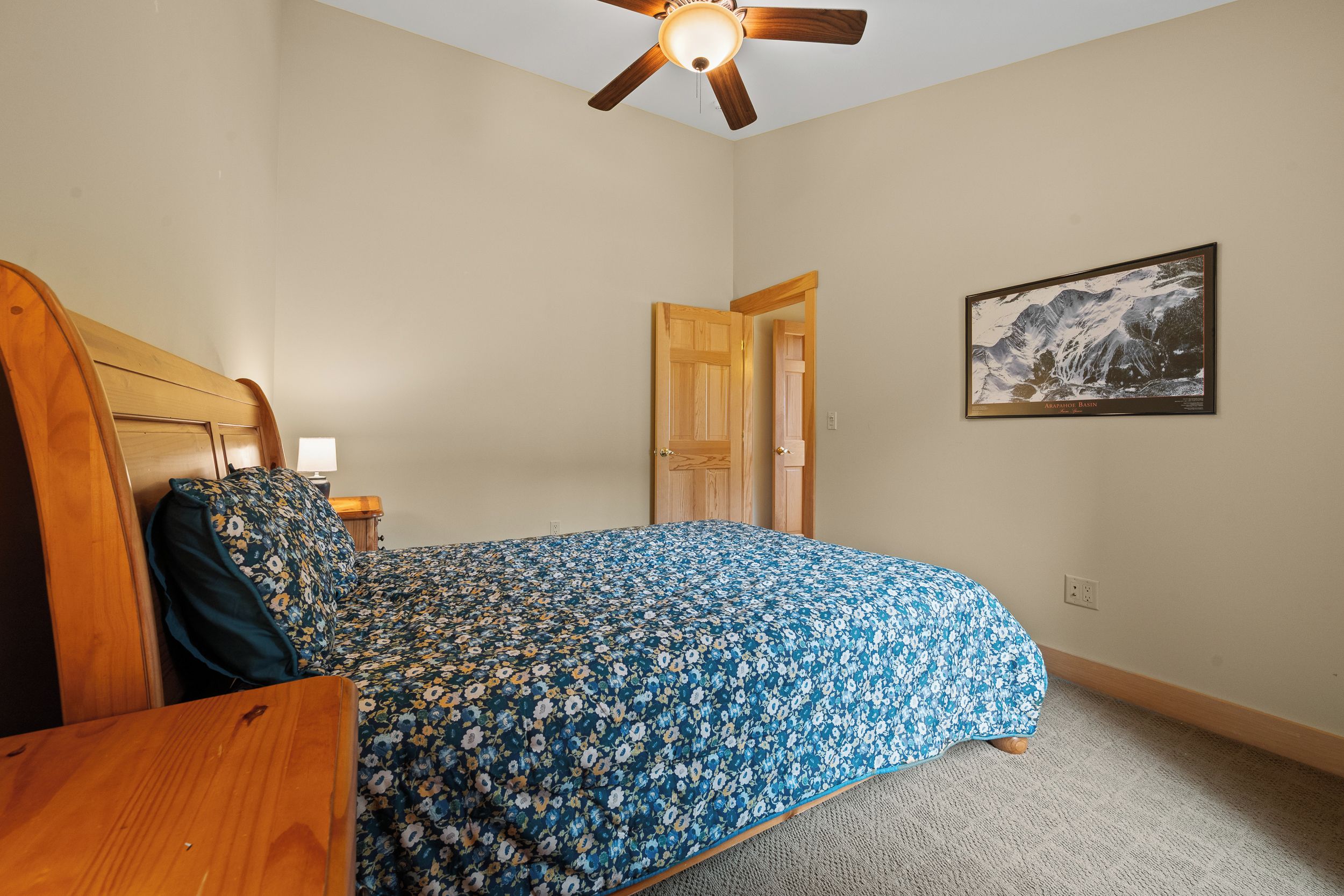 Une chambre avec un lit en bois, un ventilateur de plafond, une table de chevet, un tableau encadré au mur et un sol recouvert d’une moquette.