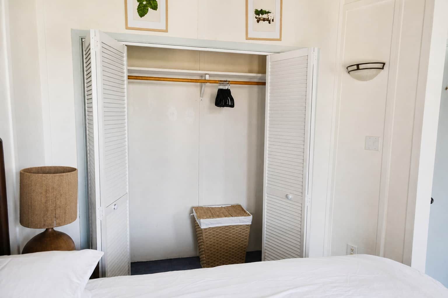 A hotel room with a bed, a wardrobe with a hanging rack, a lamp, and a small basket.