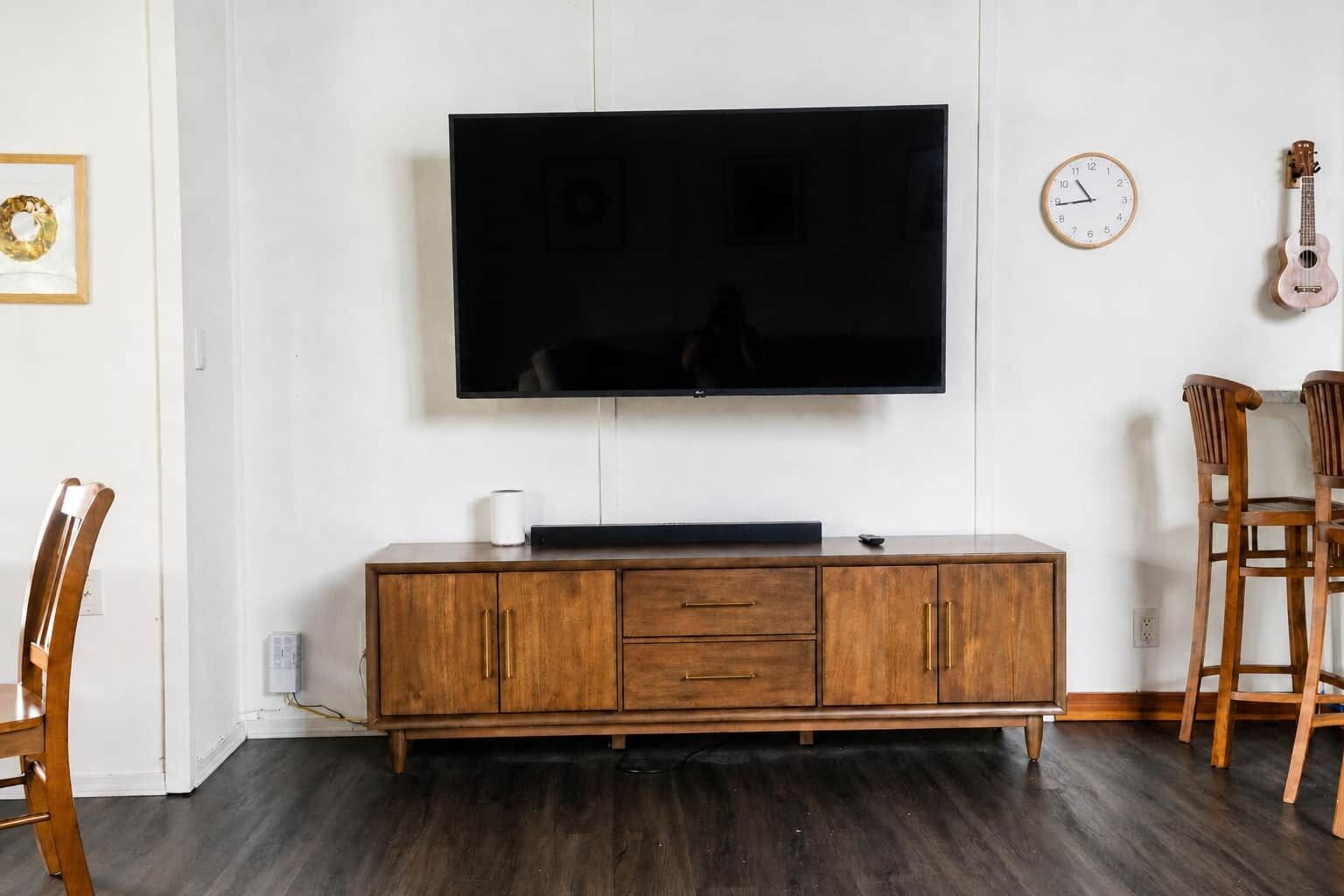 A living room with a flat-screen TV mounted on the wall, a wooden cabinet below, a clock, and two wooden chairs.