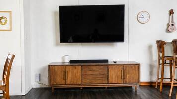 A living room with a flat-screen TV mounted on the wall, a wooden cabinet below, a clock, and two wooden chairs.