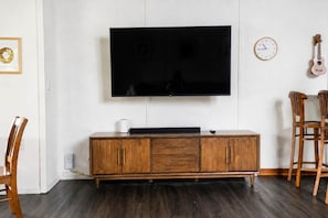 Una sala de estar con un televisor de pantalla plana montado en la pared, un mueble de madera debajo, un reloj y dos sillas de madera.
