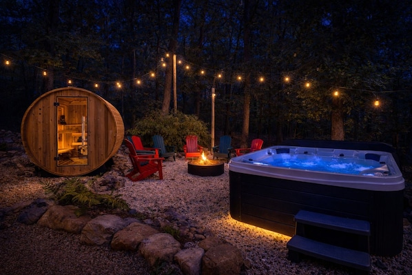 Een houten chalet met een glazen deur, buiten zitplaatsen, een jacuzzi en lichtjes in een bosrijke omgeving.
