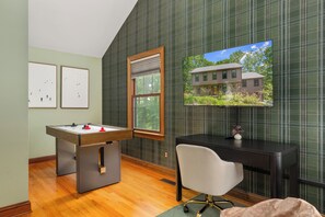 Une pièce avec un baby-foot, un bureau avec un ordinateur, une chaise et un téléviseur qui diffuse une image d’une maison.