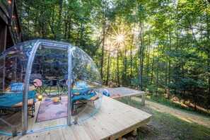 Eine transparente Kuppelstruktur mit einem Bett im Inneren, umgeben von einer Holzterrasse und einem bewaldeten Gebiet.