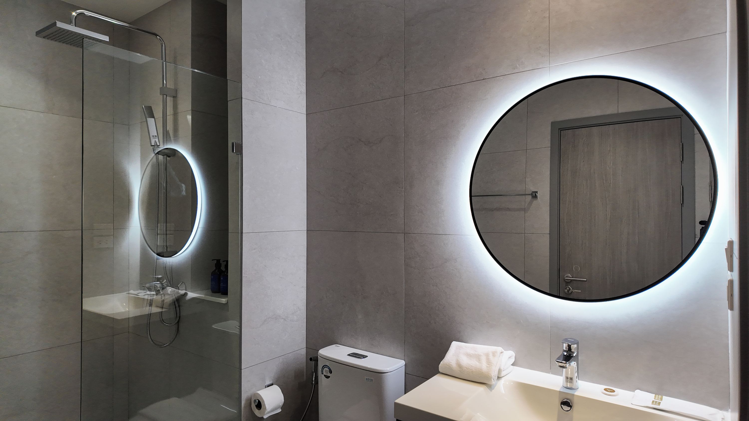 Une salle de bain moderne dotée d’une douche à parois de verre, d’un miroir rond avec éclairage intégré, de toilettes et d’un lavabo avec un robinet.