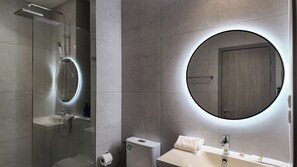 Un baño moderno con ducha envuelta en vidrio, espejo redondo con iluminación integrada, inodoro y lavabo con grifo.
