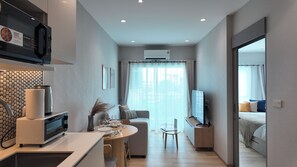 A modern kitchen with a microwave, kettle, and a coffee machine. There is a dining area with a table, chairs, and a sofa. A TV is mounted on the wall, and an air conditioning unit is visible. - Lumi 1br suite | Title Serenity Nai Yang (Tambon Sa Khu)