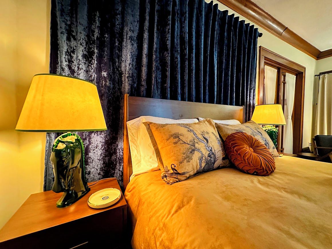 Ein Hotelzimmer mit einem großen Bett, zwei Nachttischlampen, einem Nachttisch mit einer Vase und einem dunklen Vorhang im Hintergrund.