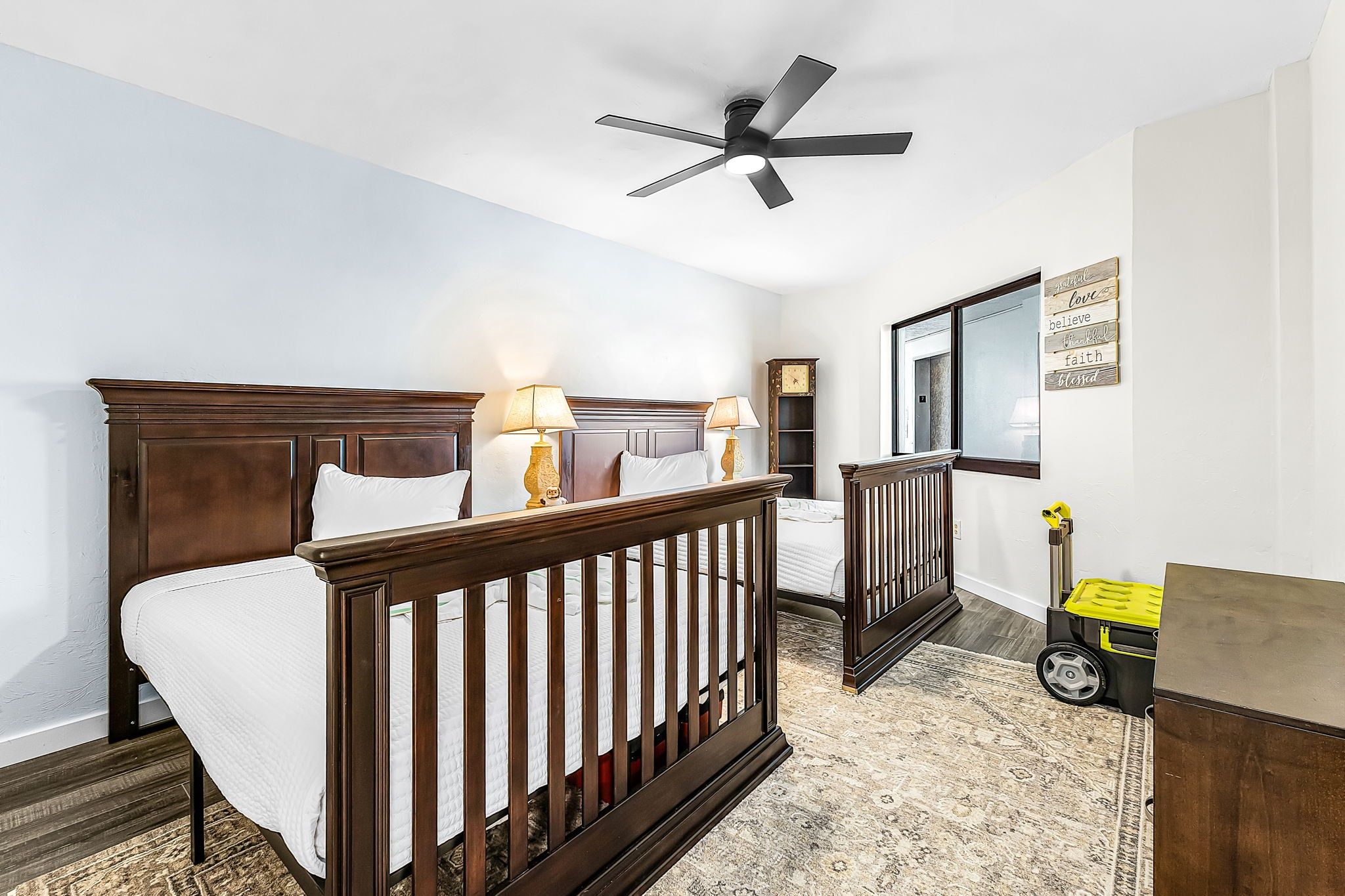Un dormitorio con una cama de madera, dos lámparas de noche, una cuna, un ventilador de techo y una frase motivacional en la pared.