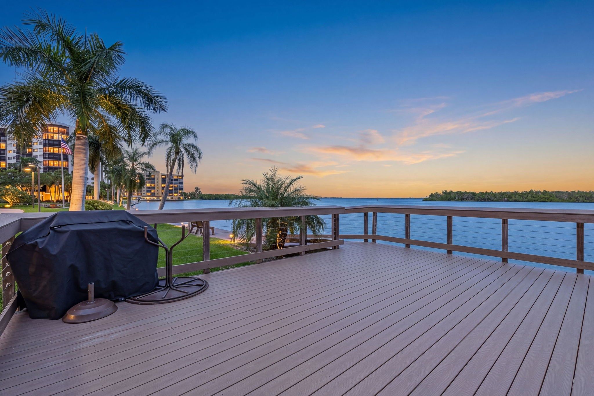 A wooden deck with a grill and outdoor furniture, overlooking a body of water with palm trees and a building in the background.