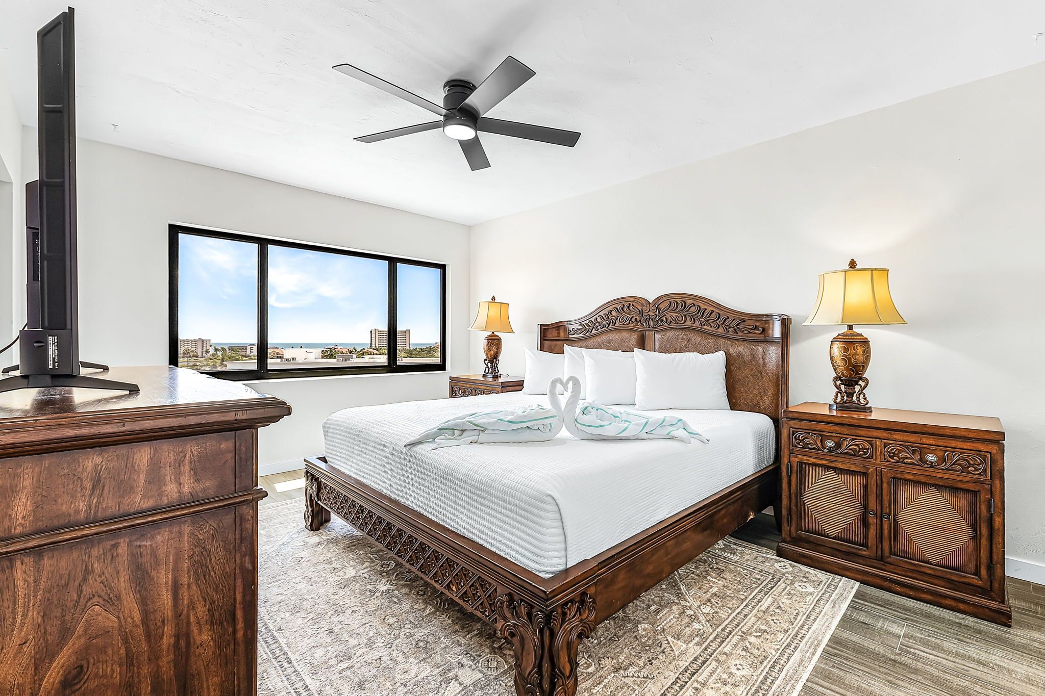 A bedroom with a large bed, a ceiling fan, a TV, and a view of the city.