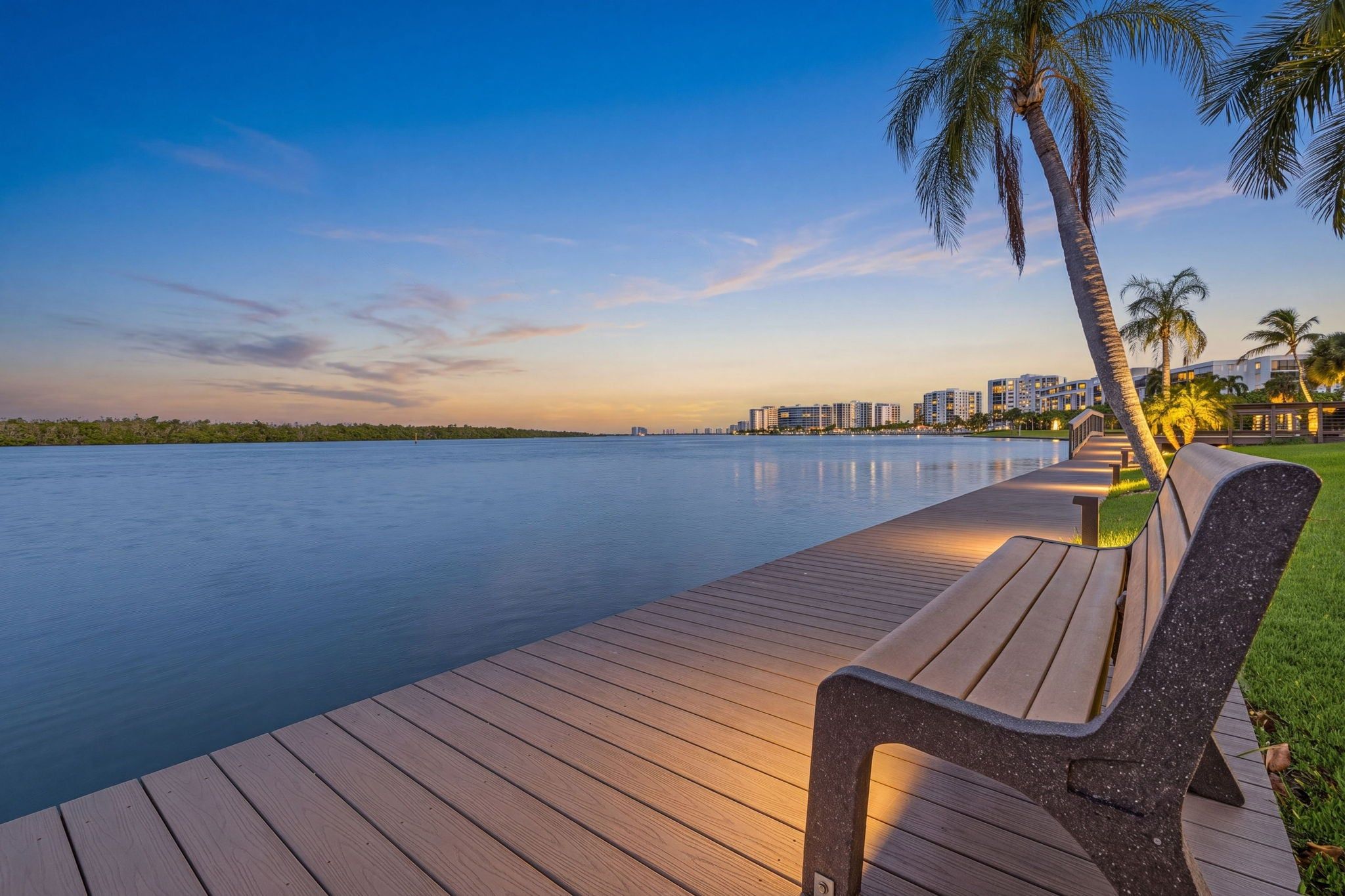 Une terrasse en bois avec un banc, des palmiers et une vue sur une étendue d’eau calme et des bâtiments situés au loin.