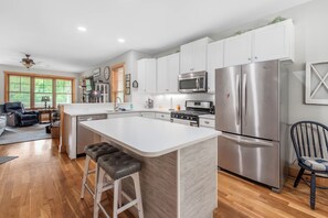 A modern kitchen with white cabinetry, stainless steel appliances, a central island with stools, and a dining area with a chair. - Mountain Gem - Minutes to Downtown WJ - Shops - Hiking (West Jefferson)
