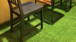 Quarto com mesa, duas cadeiras, janela com grades de metal e piso de grama artificial.