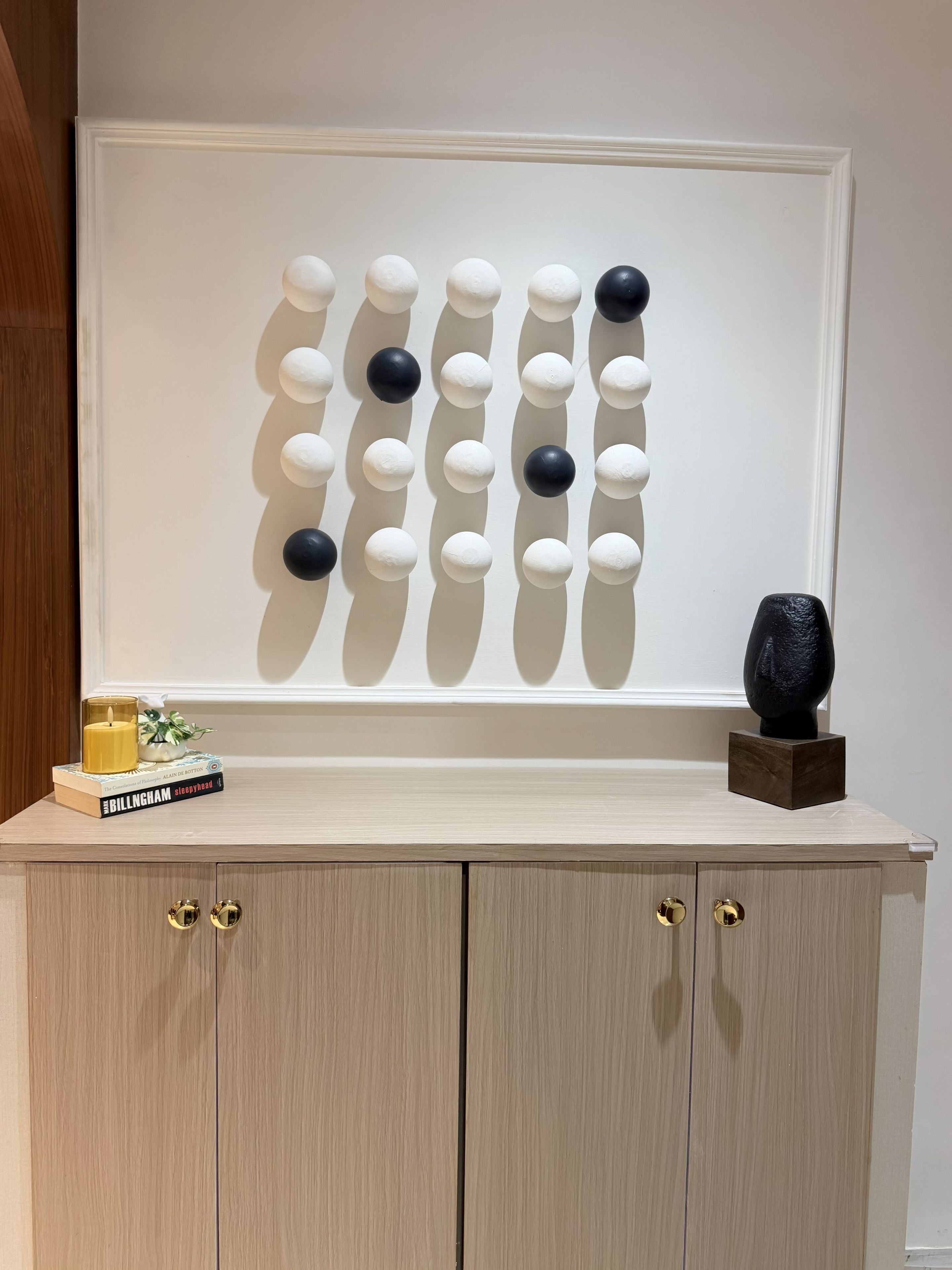 A wall-mounted artwork with a pattern of white and black spheres, a small sculpture on a wooden block, and a candle on a stack of books on a wooden cabinet.