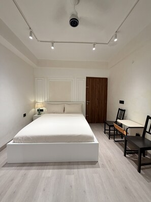 Ein Schlafzimmer mit einem großen Bett, einer Holztür, einem Schreibtisch mit Stühlen und einer an der Decke befestigten Überwachungskamera.