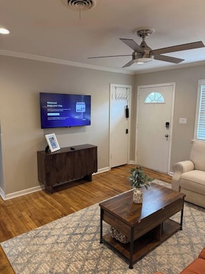 Een woonkamer met een flatscreen televisie, een plafondventilator, een houten salontafel en een beige bank.
