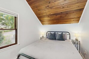A bedroom with a sloped ceiling, a bed with pillows, bedside lamps, and a window with a view of greenery. - Three Bears Cabin | Hot Tub, Pet Friendly, Minutes from Blowing Rock (Blowing Rock)