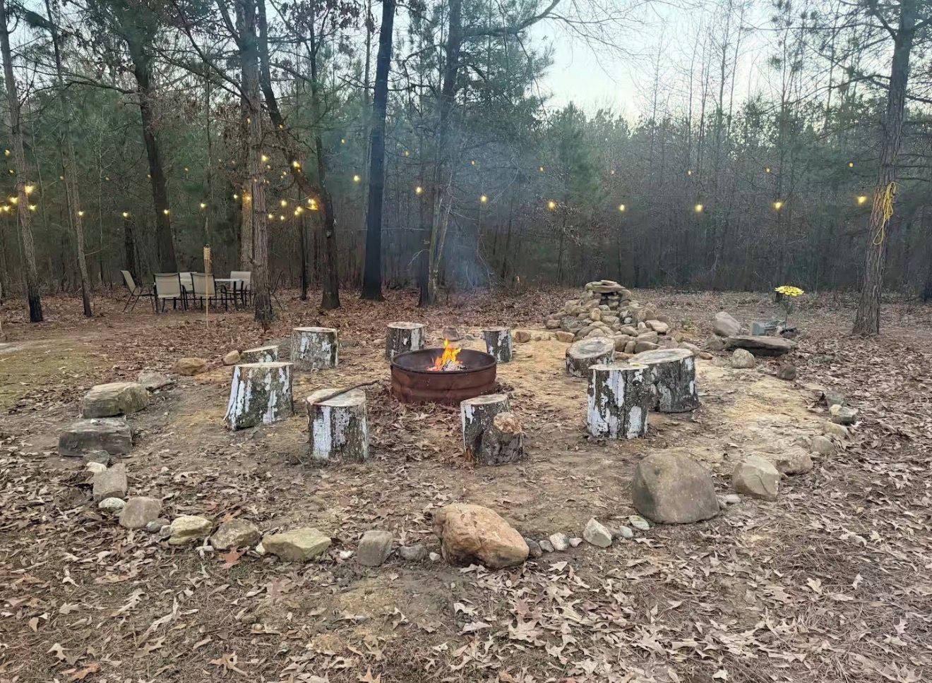 Ein Feuerkorb im Freien, umgeben von Steinen und Bäumen, mit Lichterketten darüber.