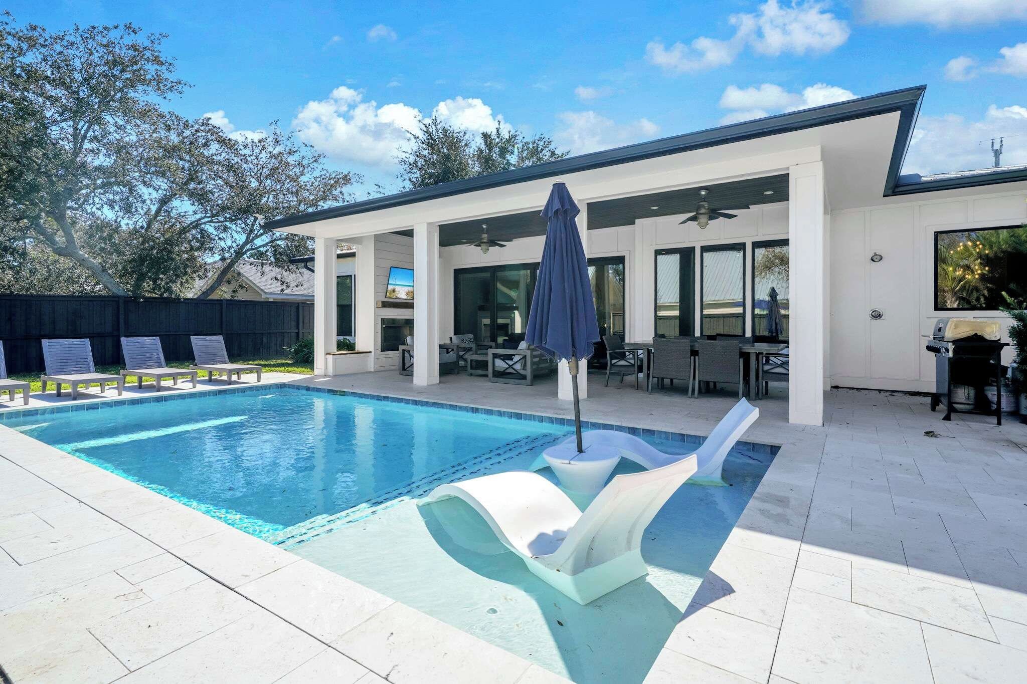 A modern outdoor swimming pool with a covered patio area, lounge chairs, and a ceiling fan.
