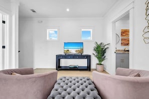 Ein Wohnzimmer mit Fernseher, zwei Fenstern, einer Zimmerpflanze und einem Sofa.