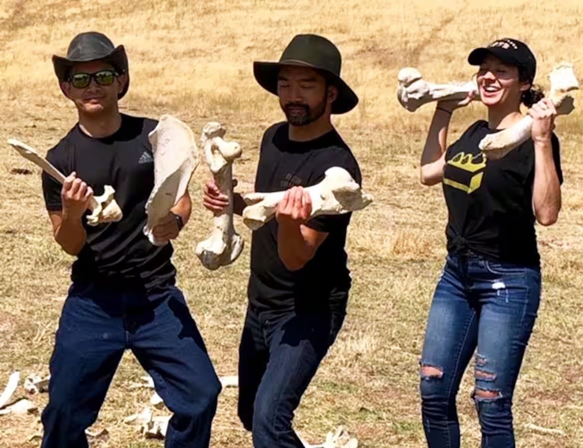 Three individuals holding large bones, standing in an open field.