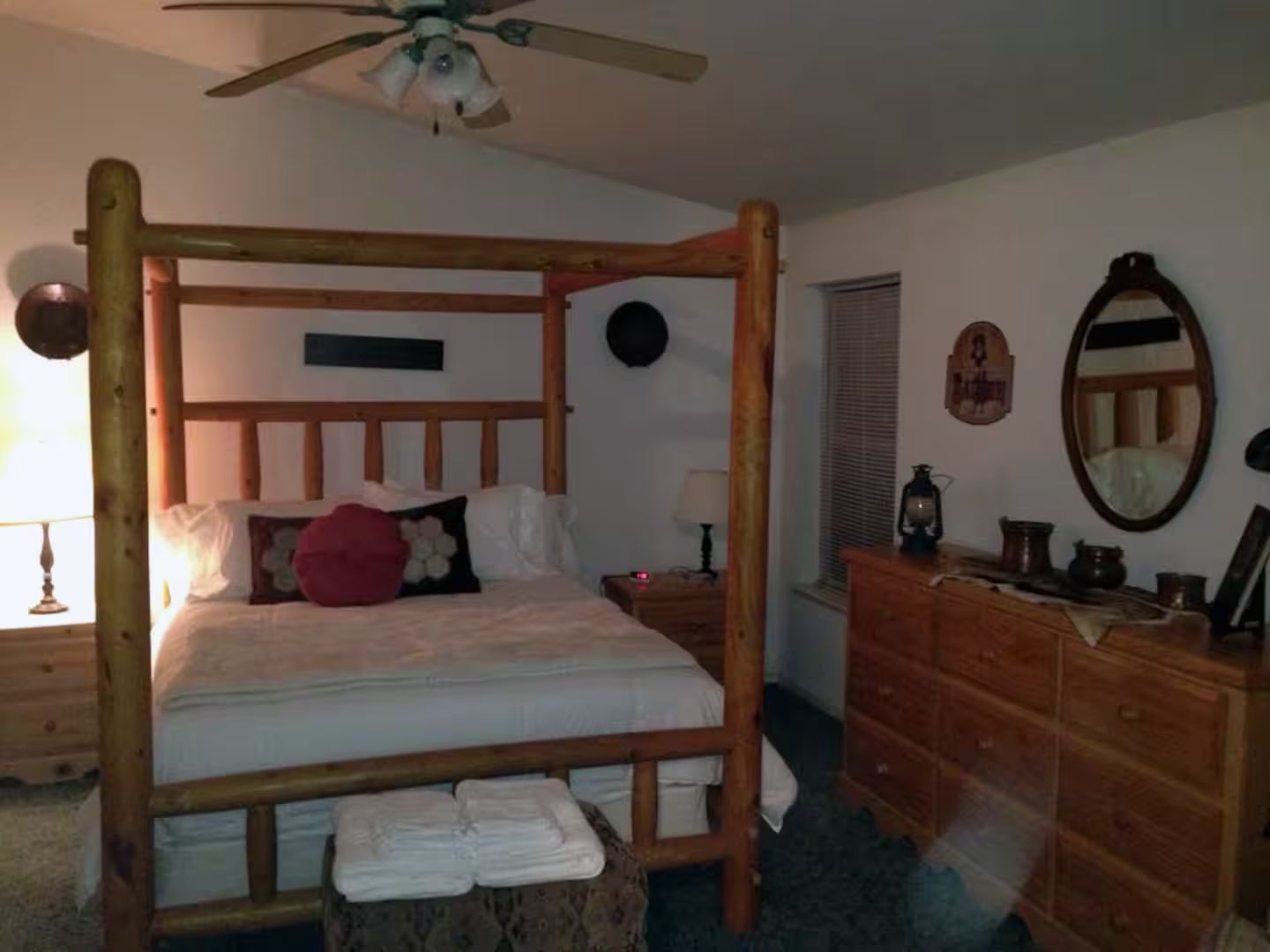 Una cama con dosel sobre un armazón de madera, un aparador de madera con cajones, un espejo redondo y un ventilador de techo.