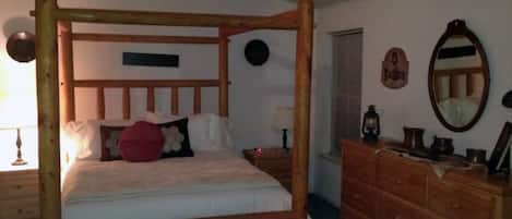 Una cama con dosel sobre un armazón de madera, un aparador de madera con cajones, un espejo redondo y un ventilador de techo.