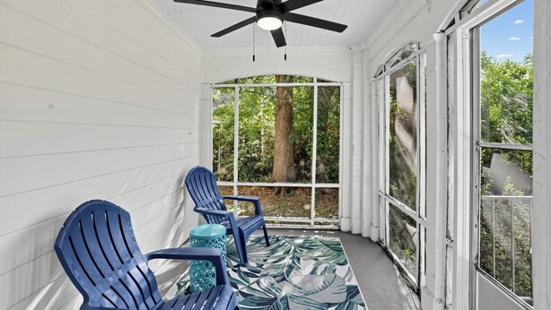 A covered porch with two blue Adirondack chairs, a ceiling fan, and a view of greenery through a window.