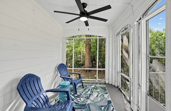 A covered porch with two blue Adirondack chairs, a ceiling fan, and a view of greenery through a window.