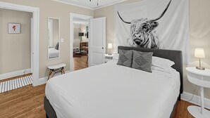 A bedroom with a large bed, a ceiling fan, a tapestry of a bull, and a small table with a lamp.