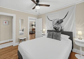 A bedroom with a large bed, a ceiling fan, a tapestry of a bull, and a small table with a lamp.