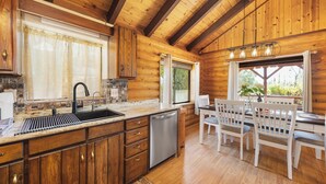 A wooden cabin interior with a kitchen, dining area, and a window with a view of greenery. - Lake and Ski/Gameroom/Dogrun (Wofford Heights)