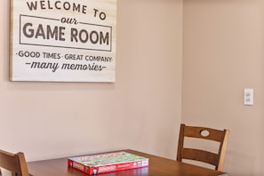 Ein Holzschild mit der Aufschrift Willkommen im Spielzimmer, ein Monopol-Spiel auf einem Tisch und zwei Holzstühle.