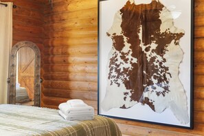 Ein Zimmer mit Holzwänden, einem Bett mit einer grün-weiß gestreiften Tagesdecke, einem Stapel weißer Handtücher und einem gerahmten Tierfell an der Wand.