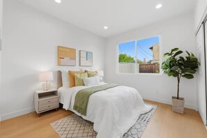 Quarto com cama, mesas de cabeceira, um criado-mudo, janela e planta em vaso.