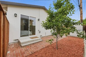 Edifício residencial com entrada por porta de vidro deslizante, uma pequena árvore no jardim e um caminho de tijolos até a porta.