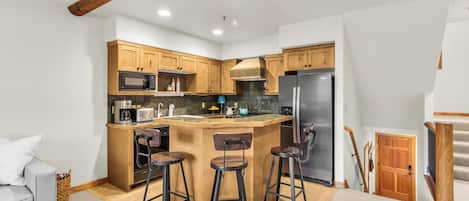 A compact kitchen with wooden cabinets, a built-in microwave, a stainless steel refrigerator, and a bar stool area.