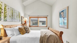Ein Schlafzimmer mit einem Bett, einem Nachttisch, einer Lampe, einem Fenster mit Jalousien und Bildern an den Wänden.
