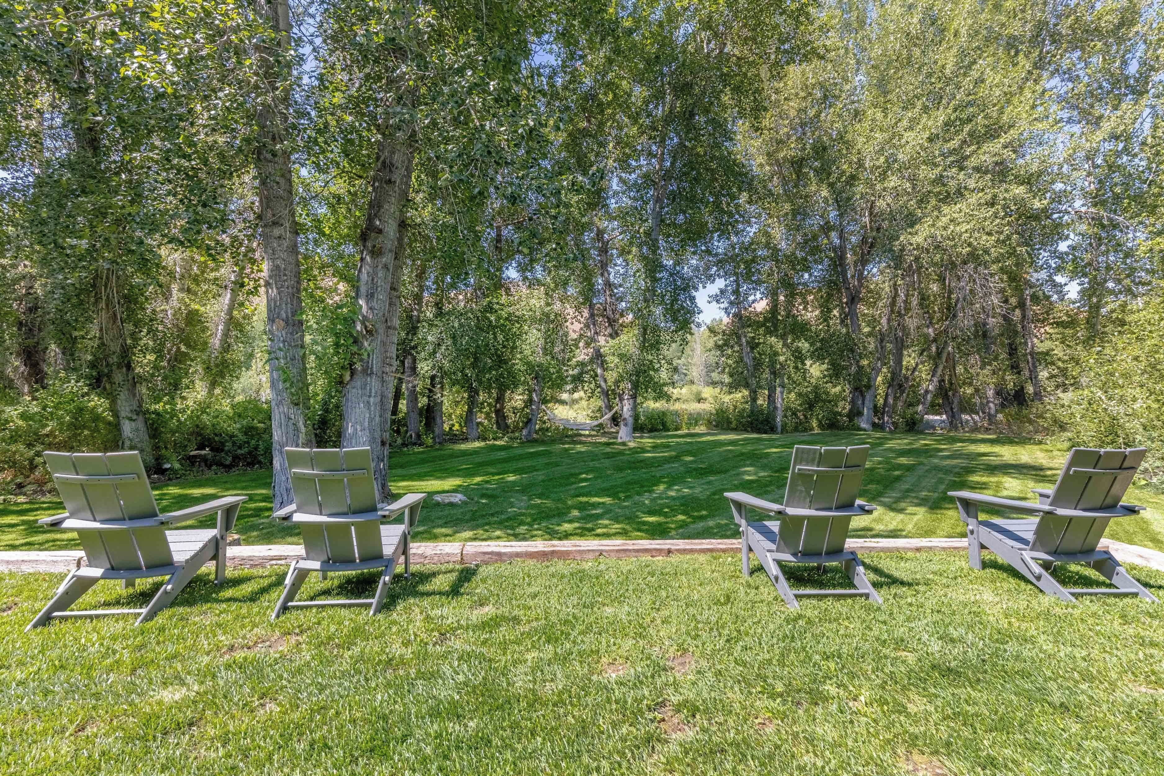 A park with four Adirondack chairs arranged in a row on a well-maintained lawn, surrounded by tall trees.
