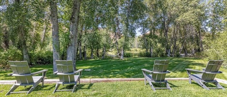 A park with four Adirondack chairs arranged in a row on a well-maintained lawn, surrounded by tall trees.