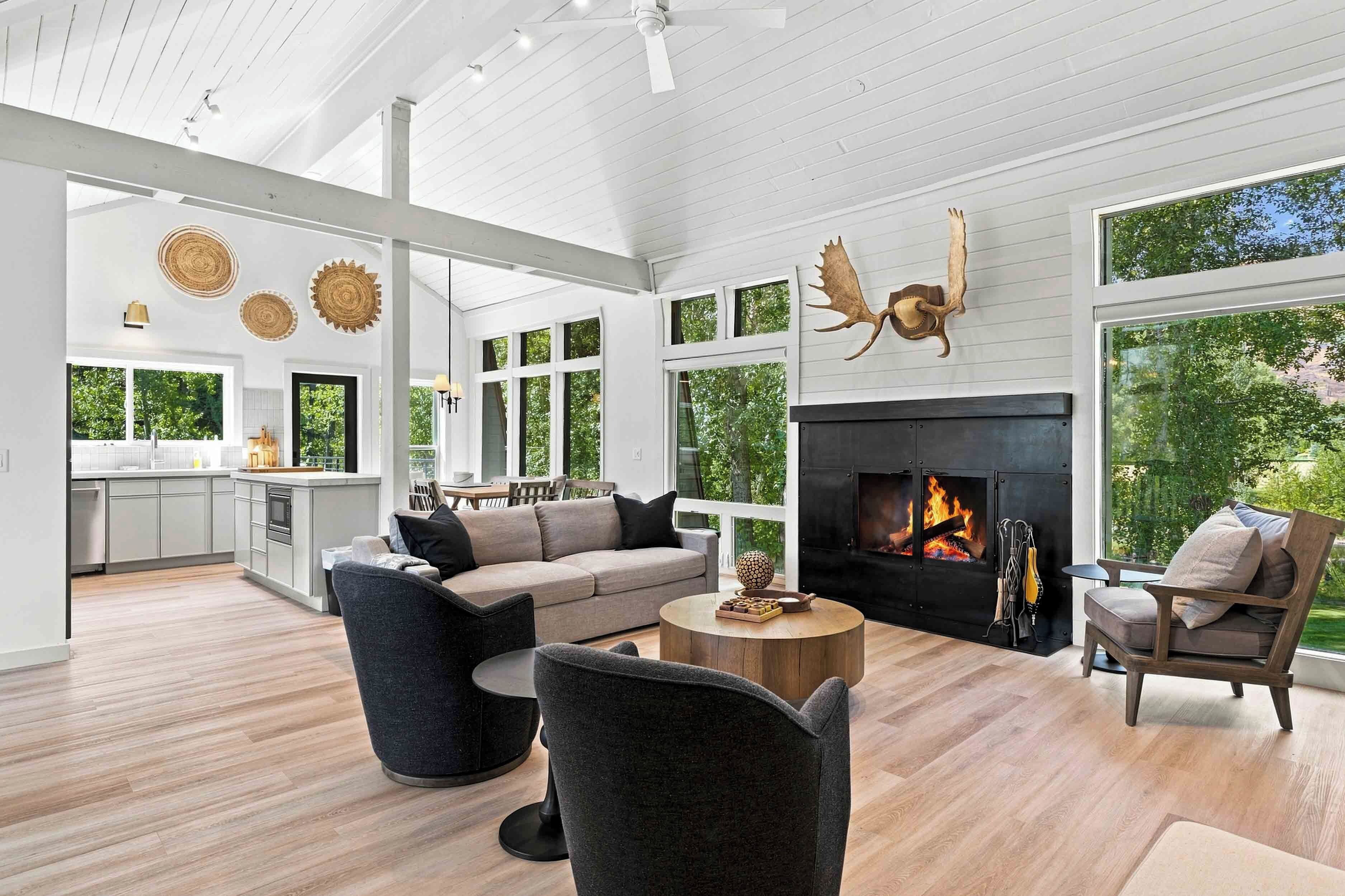 A spacious living area with a fireplace, large windows, and a modern kitchen in the background.