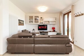 A living room with a brown leather sofa, a flat-screen TV displaying 'NETFLIX', and a kitchen area with cabinets and a window. - Villa con piscina privata tra natura e 5 terre. (Sarzana)