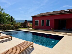 A swimming pool with wooden loungers, a red building with shutters, and a mountain view in the background. - Villa con piscina privata tra natura e 5 terre. (Sarzana)