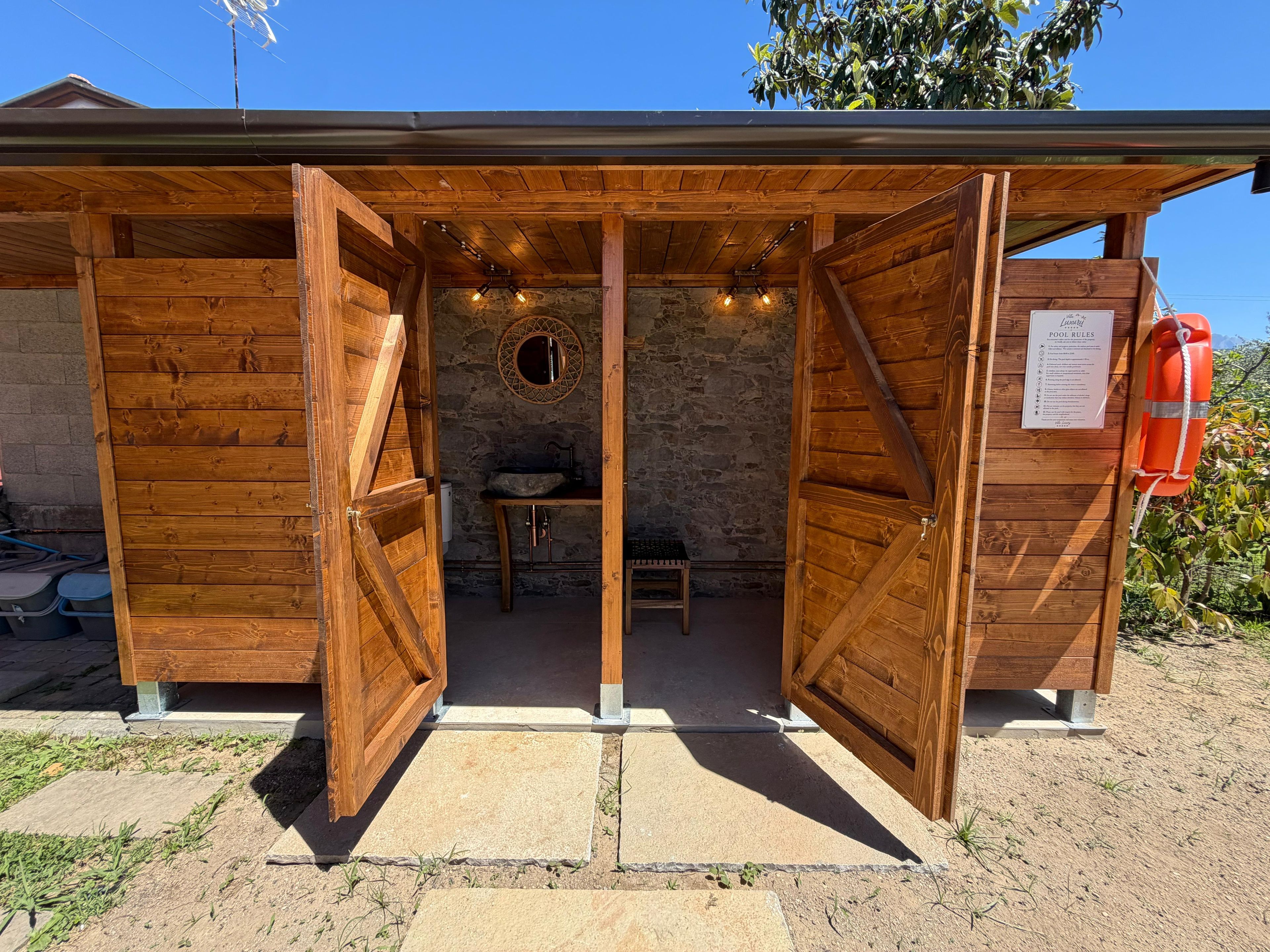 A wooden structure with open doors, a stone wall, a round mirror, a sink, and a lifebuoy.
