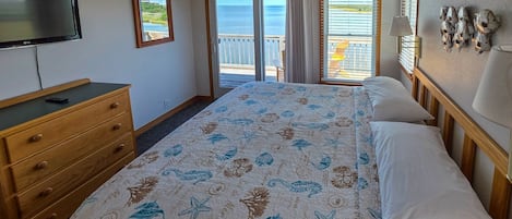 A bedroom with a bed, a dresser, a TV, a balcony with a view of the sea, and a wall with decorative items.