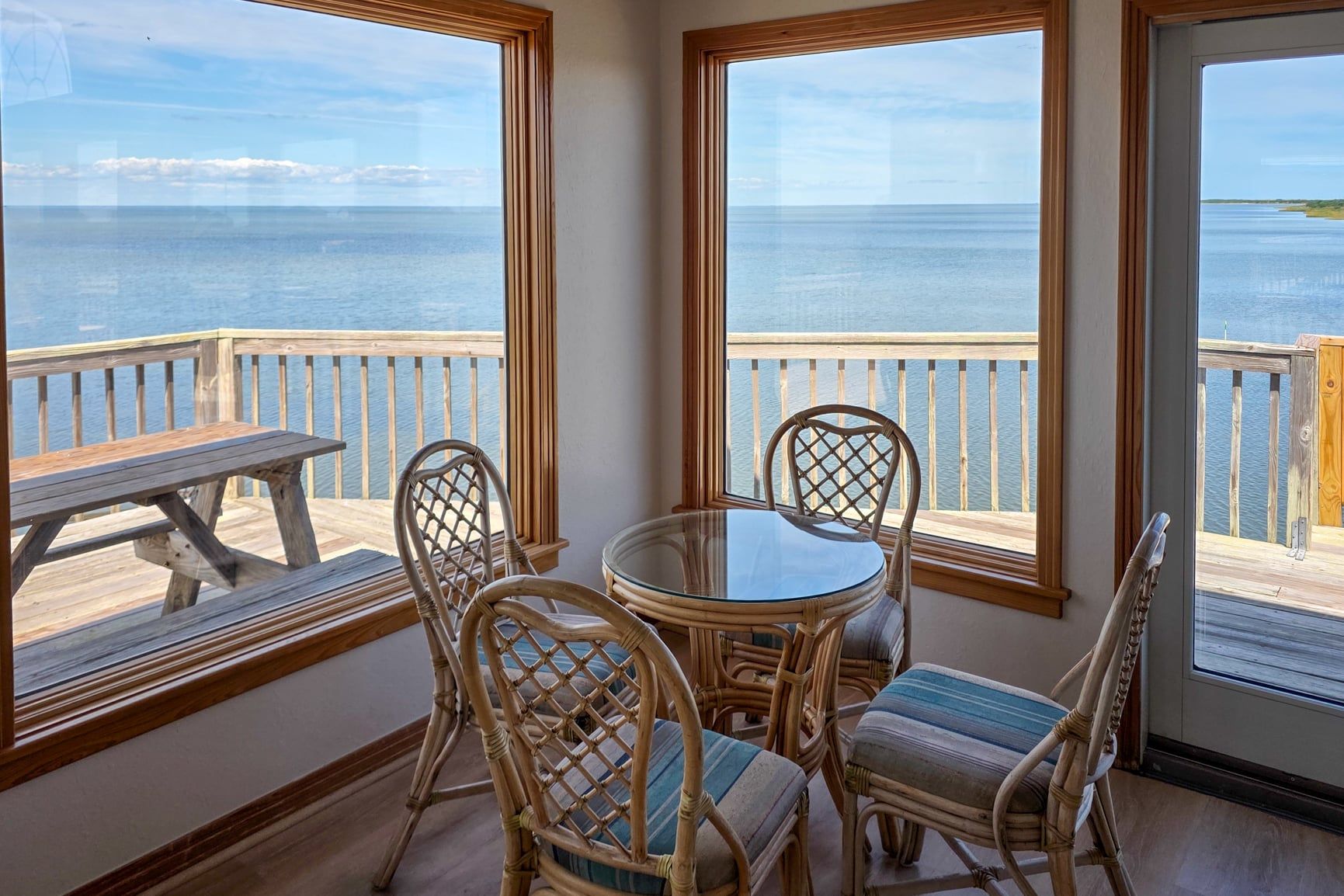 A room with a view of the sea, a round table with chairs, and a wooden deck outside.