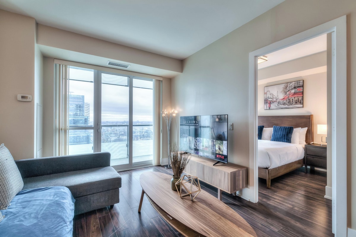 A modern hotel room with a large window, a flat-screen TV, a bed with blue pillows, and a wooden coffee table.