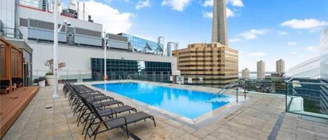 Una piscina sul tetto con lettini, circondata da edifici moderni e un cielo sereno.