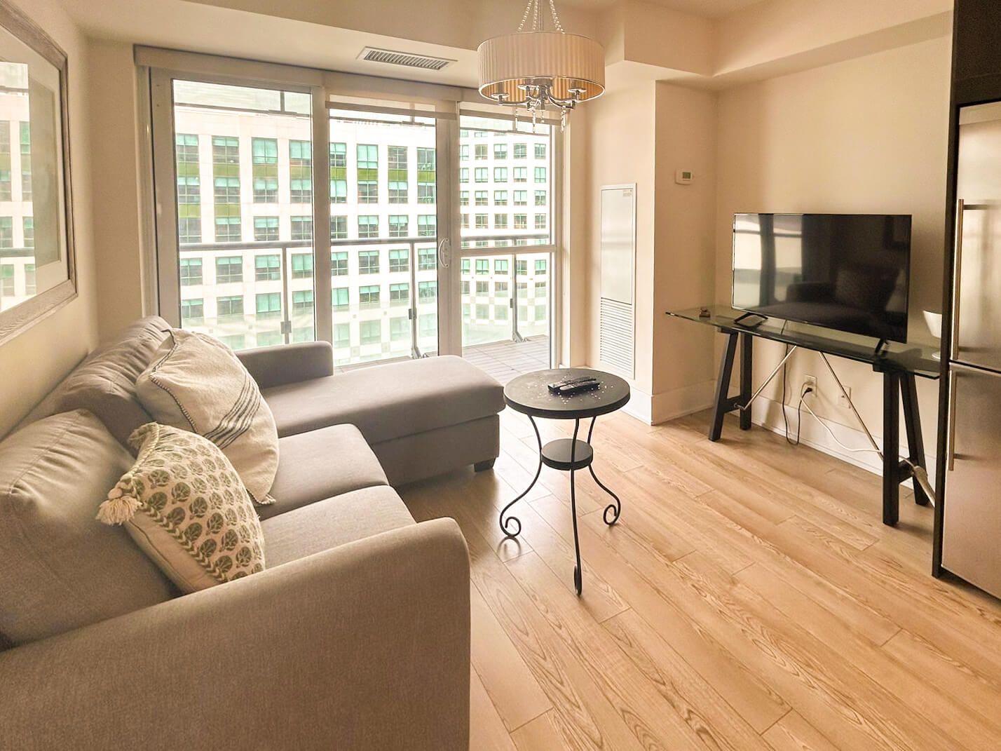 A modern living room with a sofa, a small round table, a TV on a glass stand, and large windows with a city view.